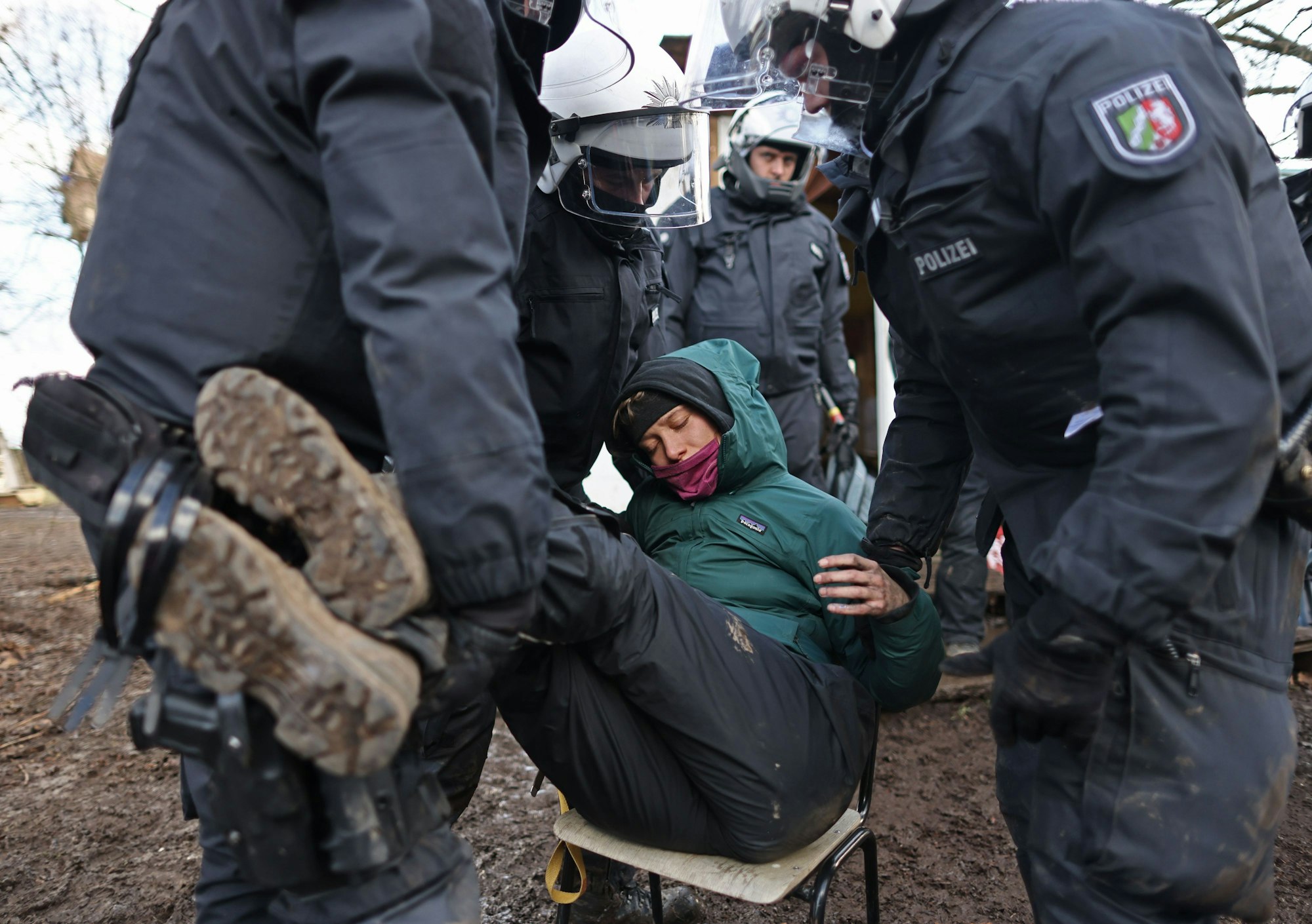 Drei Polizisten greifen eine Demonstrantin, die auf einem Stuhl sitzt, an den Beinen und Armen, um sie wegzutragen.