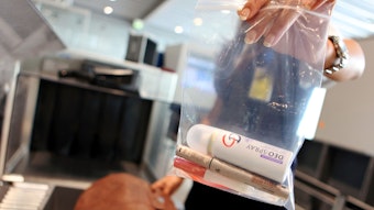 Eine Sicherheitsmitarbeiterin des Flughafens Köln/Bonn hält einen Beutel mit Kosmetika und einem Deodorant in die Kamera. Sie steht vor einer Sicherheitskontrolle. (Archivbild)