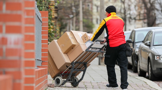 Ein DHL-Paketzusteller geht mit einer Sackkarre voller Pakete zu einem Haus. 