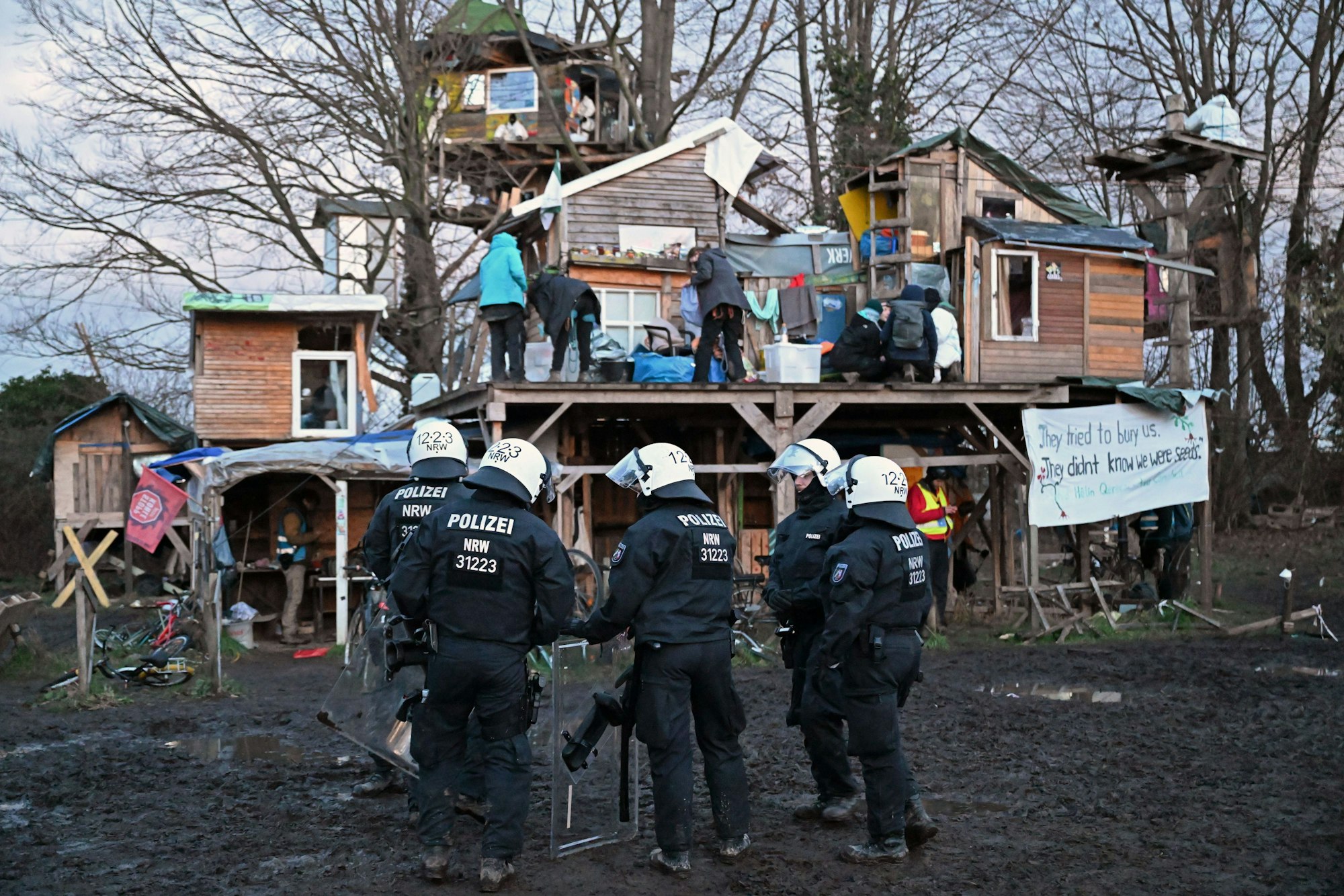 Polizisten mit Ausrüstung und Helmen stehen in einer Gruppe vor Bretterhütten, die Aktivisten aufgebaut haben. Auf einem Podest an den Hütten befinden sich mehrere Aktivisten.