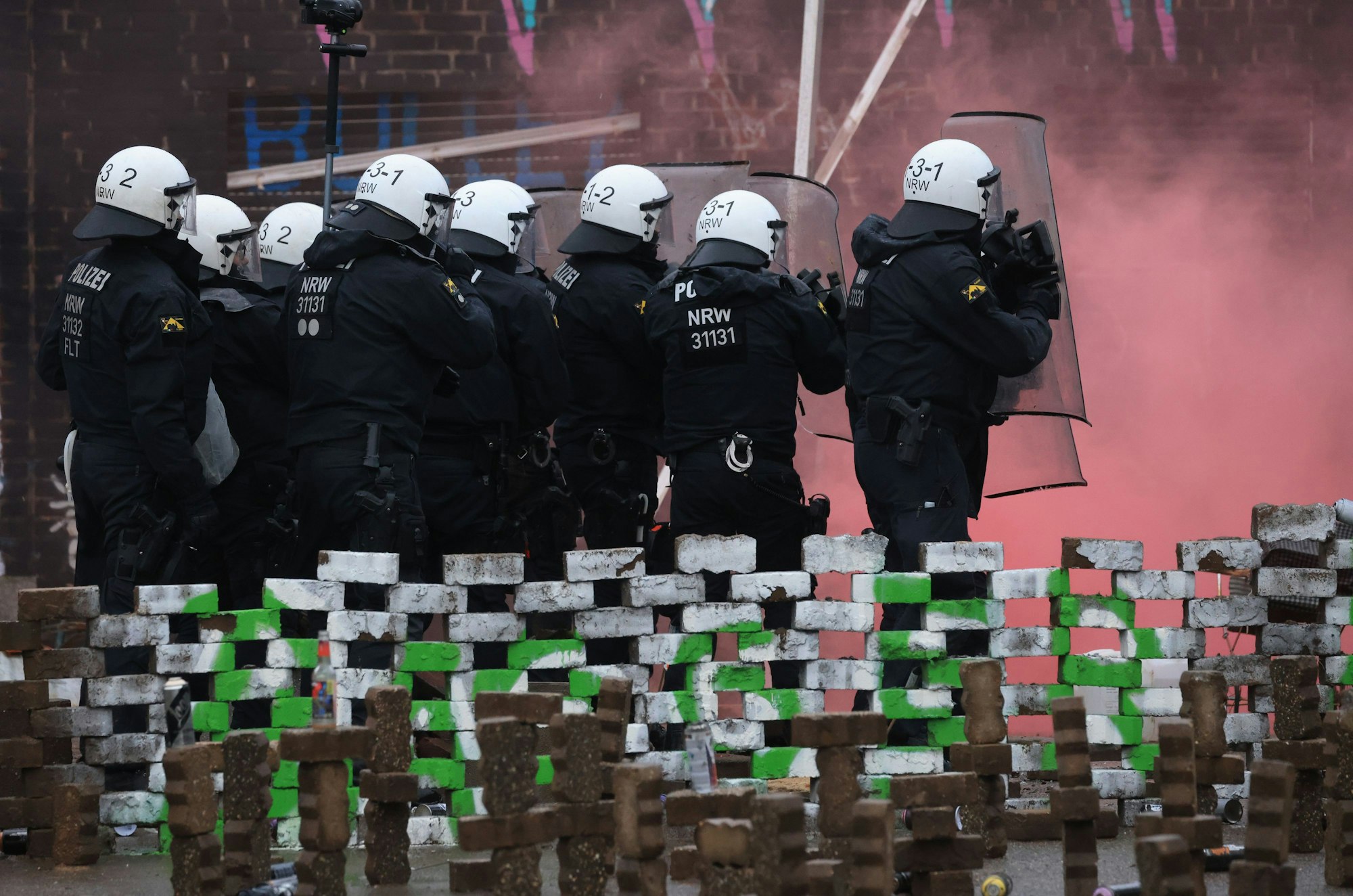 Polizisten in Schutzmontur stehen hinter Barrikaden der Klimaschützern. Vor ihnen hat sich, vermutlich durch Böller, roter Rauch gebildet.