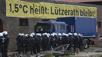 Ein Banner mit der Aufschrift "1,5°C heißt: Lützerath bleibt!" hängt an einem verlassenen Gebäude. Davor stehen Polizisten.