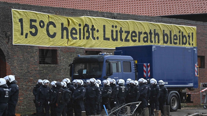 Ein Banner mit der Aufschrift "1,5°C heißt: Lützerath bleibt!" hängt an einem verlassenen Gebäude. Davor stehen Polizisten.