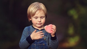Ein kleiner Junge mit gestreiftem T-Shirt und Latzhose hält ein rosafarbenes Herz in der Hand.