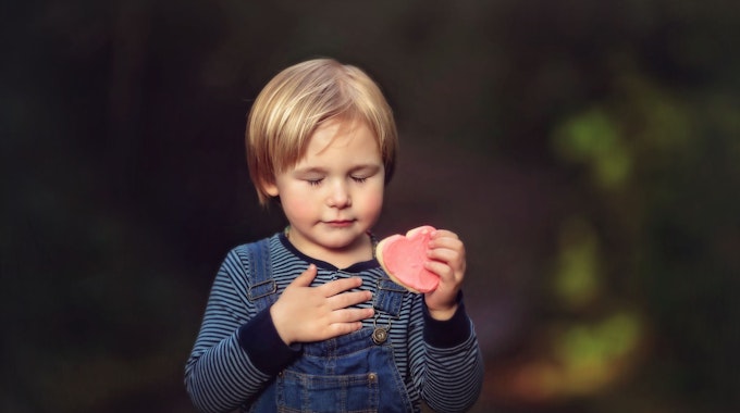 Ein kleiner Junge mit gestreiftem T-Shirt und Latzhose hält ein rosafarbenes Herz in der Hand.