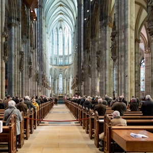 Menschen sitzen in den Bänken des Kölner Doms. Der Blick vom Foto aus geht genau mittig durch die Reihen auf den Altar.