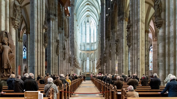Menschen sitzen in den Bänken des Kölner Doms. Der Blick vom Foto aus geht genau mittig durch die Reihen auf den Altar.