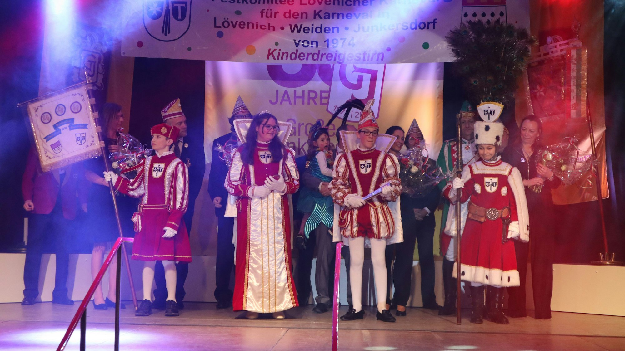 Vier Kinder in rot-weißen Karnevalstrachten stehen auf der Bühne. Hinter ihnen sind noch einige Erwachsene im Kostüm zu sehen.