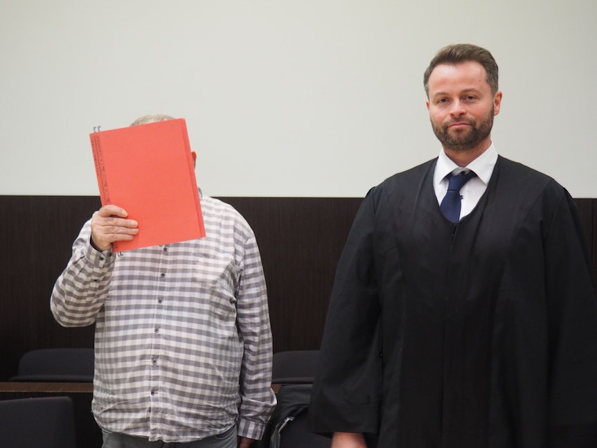 Ein Angeklagter hält sich einen roten Ordner vors Gesicht. Rechts steht sein Anwalt.