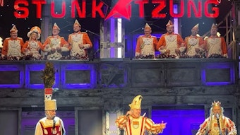 Drei Männer in Karnevalsuniform auf der Bühne, auf der Empore acht weitere Menschen in Uniform.