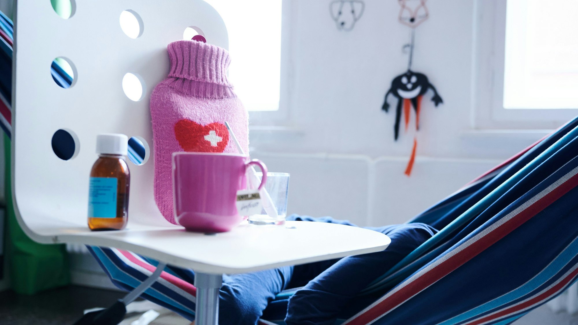 Fieberthermometer, Hustensaft, Wärmflasche und eine Tasse Tee stehen auf einem Stuhl, während ein Kind im Hintergrund in einer Hängematte liegt (Gestellte Szene).