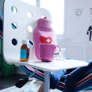 Fieberthermometer, Hustensaft, Wärmflasche und eine Tasse Tee stehen auf einem Stuhl, während ein Kind im Hintergrund in einer Hängematte liegt (Gestellte Szene).&nbsp;