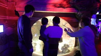 Drei Personen stehen vor einem Aquarium, eine Frau hält ihr Kind auf dem Arm. Der Raum ist in rotes und violettes Licht getaucht.