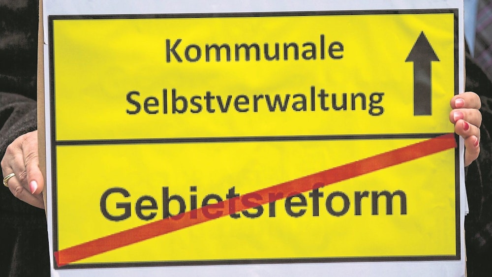 Ein gelbes Schild: Oben die Aufschrift „Kommunale Selbstverwaltung“ mit einem Pfeil nach oben daneben, unten die Aufschrift „Gebietsreform“ mit einem roten Balken durchgestrichen.