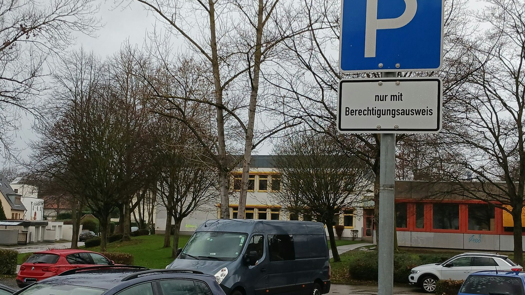 Einige Fahrzeuge parken auf einem asphaltierten Platz, hinter dem kahle Bäume und das graue Flachdachgebäude des Schulzentrums Neuenhof stehen.