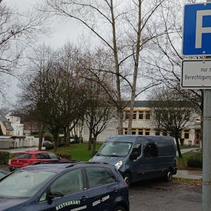 Einige Fahrzeuge parken auf einem asphaltierten Platz, hinter dem kahle Bäume und das graue Flachdachgebäude des Schulzentrums Neuenhof stehen.&nbsp;