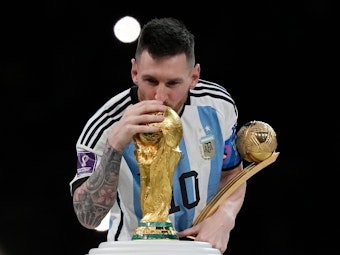 Lionel Messi küsst den WM-Pokal und hält dabei seine Trophäe als bester Spieler der WM 2022 in der Hand.