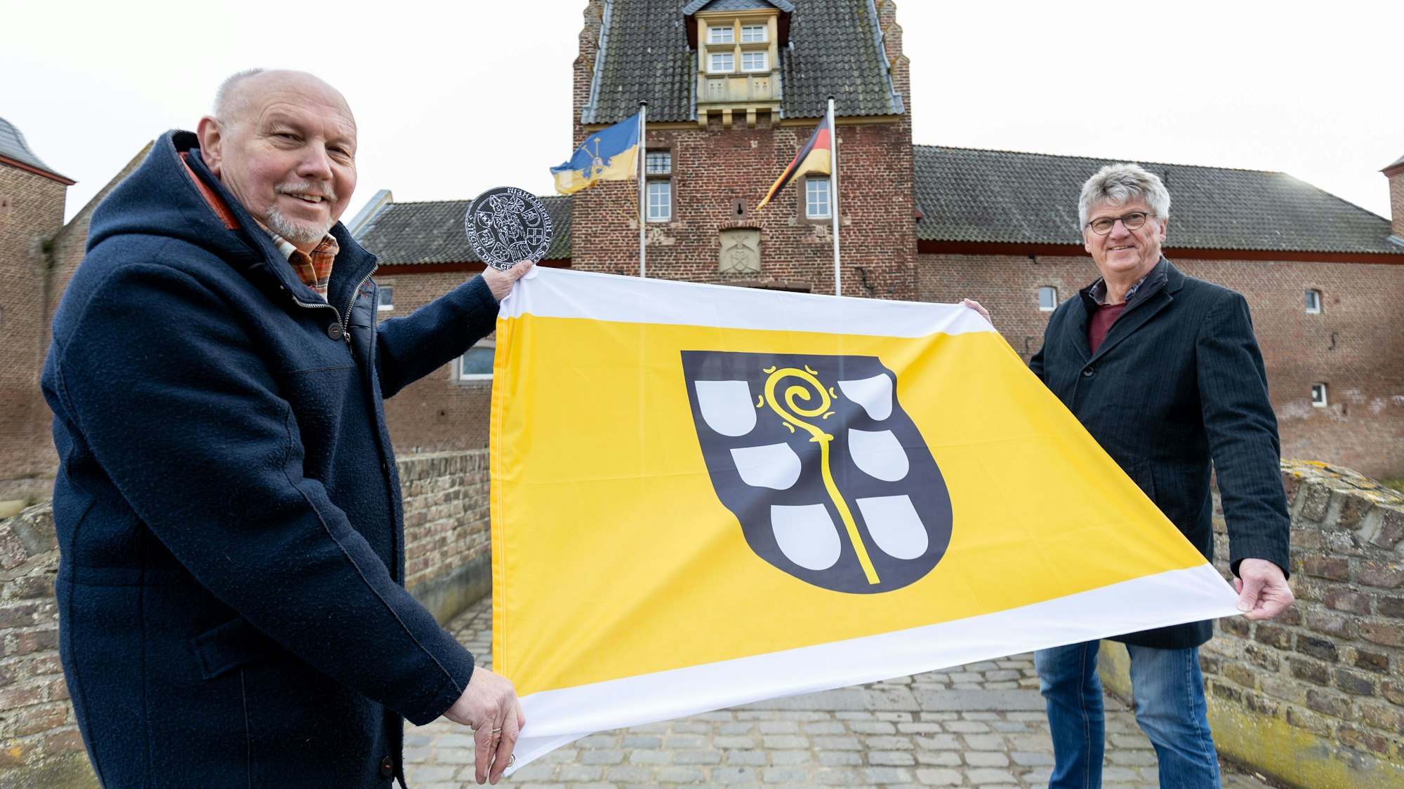 Der ehemalige Ortsvorsteher Rainer Schmidt und der aktuelle Ortsvorsteher Herrmann Menth zeigen die neue, jetzt bestellbare Fahne Heimerzheims vor dem Schloss Heimerzheim am 10. Januar 2023.