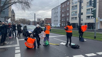 Ein Mann mit grauer Jacke greift Klimaaktivisten mit orangen Warnwesten bei einer Straßensperre in der Kölner Innenstadt an.