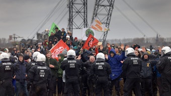 Polizisten und Demonstranten mit erhobenen Händen stehen sich bei der Demonstration von Klimaaktivisten am Rande des Braunkohletagebaus bei Lützerath gegenüber, im Hintergrund ein Kohlebagger.