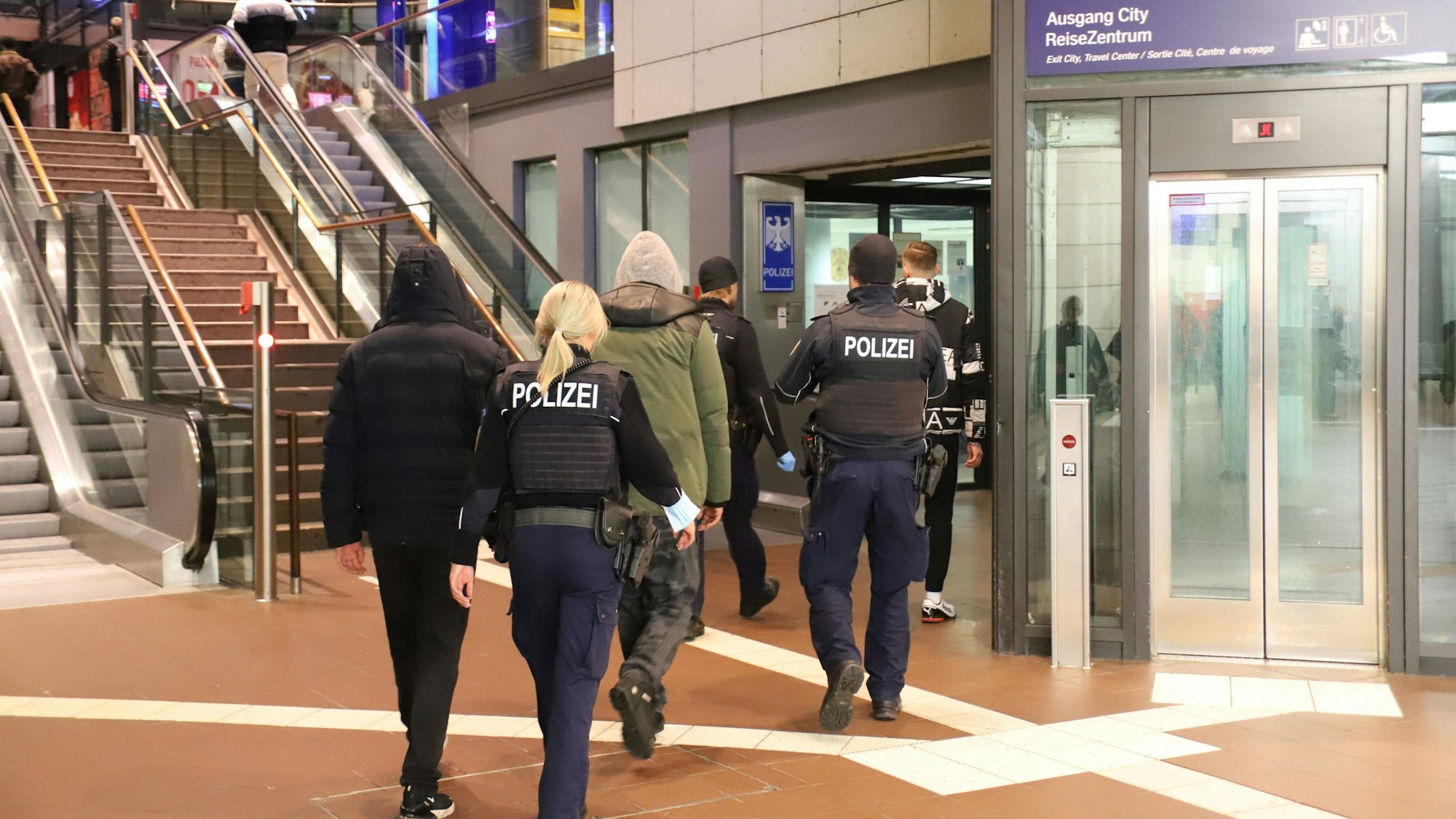 Im Siegburger Bahnhof vor den Treppen und Rolltreppen und neben dem Aufzug gehen zwei uniformierte Beamte. Sie sind von hinten zu sehen, eine blonde Frau mit Pferdeschwanz und ein Beamter mit Mütze begleiten drei Männer, die Kapuzenjacken tragen.