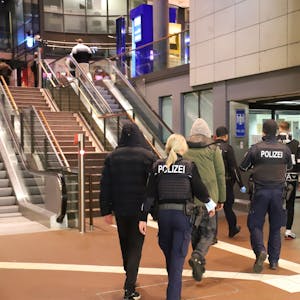 Im Siegburger Bahnhof vor den Treppen und Rolltreppen und neben dem Aufzug gehen zwei uniformierte Beamte. Sie sind von hinten zu sehen, eine blonde Frau mit Pferdeschwanz und ein Beamter mit Mütze begleiten drei Männer, die Kapuzenjacken tragen.