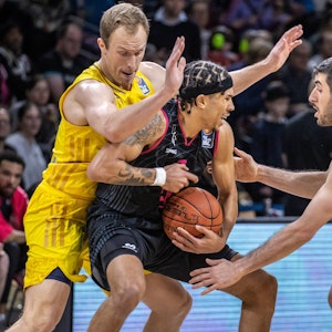 Zwei Basketballer in gelben Trainingsanzügen versuchen einem gegenerischen Spieler den Ball abzunehmen.