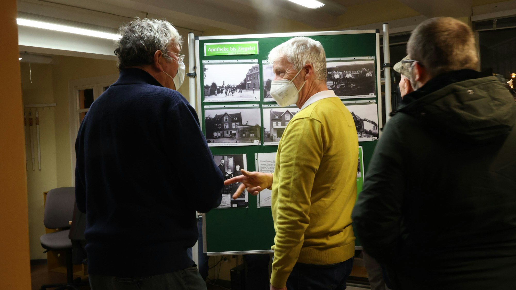 Vor einer Tafel mit historischen Fotos unterhalten sich zwei ältere Männer.