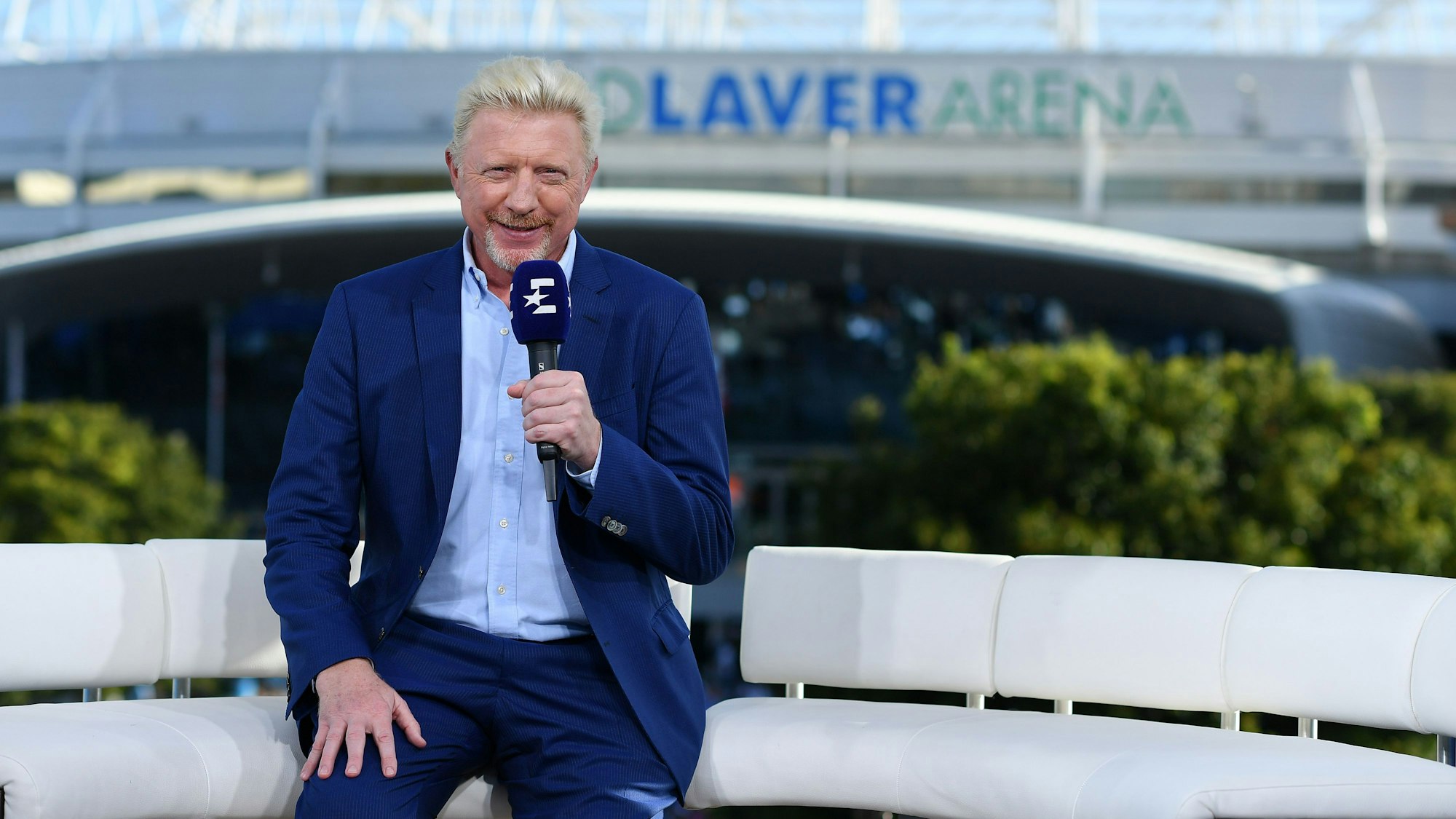 Boris Becker sitzt mit einem Eurosport-Mikrofon in der Hand auf einem Sofa.
