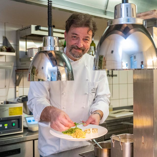 Roberto Carturan steht in&nbsp;&nbsp;Kochkleidung in seiner Küche und hält einen Teller mit Essen in der Hand