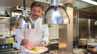 Roberto Carturan steht inKochkleidung in seiner Küche und hält einen Teller mit Essen in der Hand