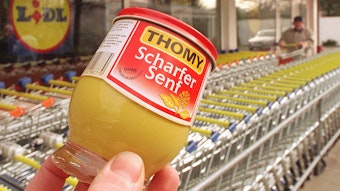 Vor abgestellten Einkaufswagen deines Lidl-Lebensmittelgeschäftes wird symbolisch ein Senfglas der Firma Thomy ins Bild gehalten.