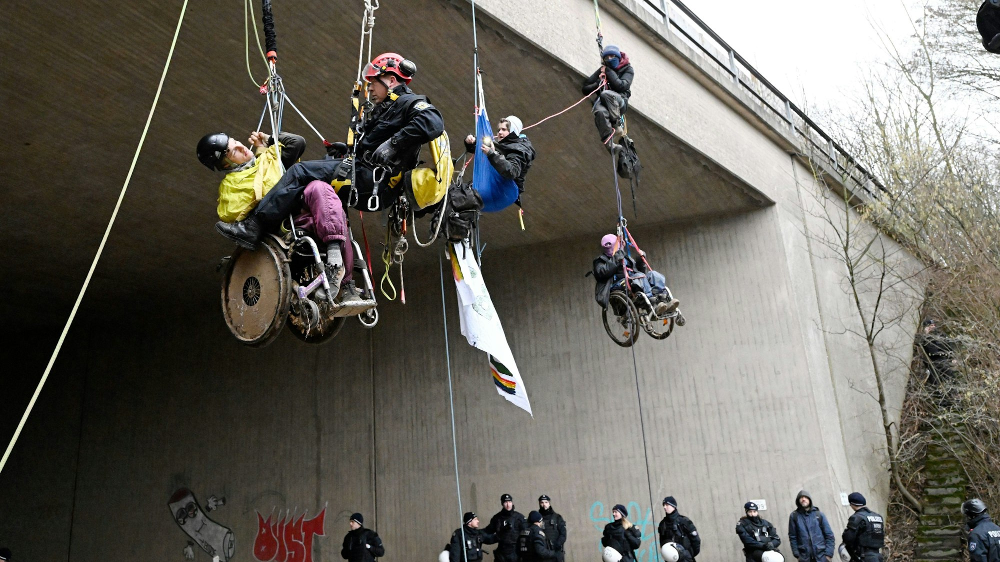 Ein Höhenretter der Polizei hilft einem  Umweltaktivisten im Rollstuhl auf den Boden. Klimaaktivisten in Rollstühlen haben sich am Montagmorgen von einer Autobahnbrücke der A44 in der Nähe von Lützerath abgeseilt. Die Brücke liegt ungefähr vier Kilometer Luftlinie vom Braunkohleort Lützerath entfernt.
