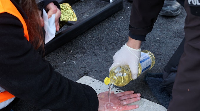 Eine an einer Fahrbahnmarkierung festgeklebte Hand wird von einem Rettungsdienstmitarbeiter mit Speiseöl übergossen.