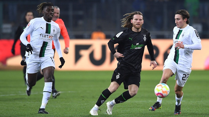 Rocco Reitz (r.) und Manu Koné (l.) treiben den Ball im weißen Heimtrikot von Borussia Mönchengladbach beim Legendenspiel am 17. Dezember 2022 nach vorne. Angegriffen wird Reitz von U23-Trainer Eugen Polanski (M.) im schwarzen Trikot der Borussia-Legenden.