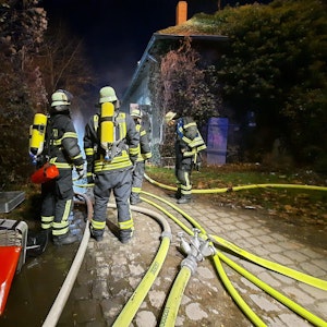 Eine Gruppe von Feuerwehrleuten mit schwerem Atemschutz stehen vor dem Gebäude; auf dem Boden liegen mehrere gelbe Löschschläuche.