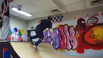 Schüler, der mit Helm und Schonern auf der Skateboard-Rampe fährt, während ein weiterer Schüler zuguckt
