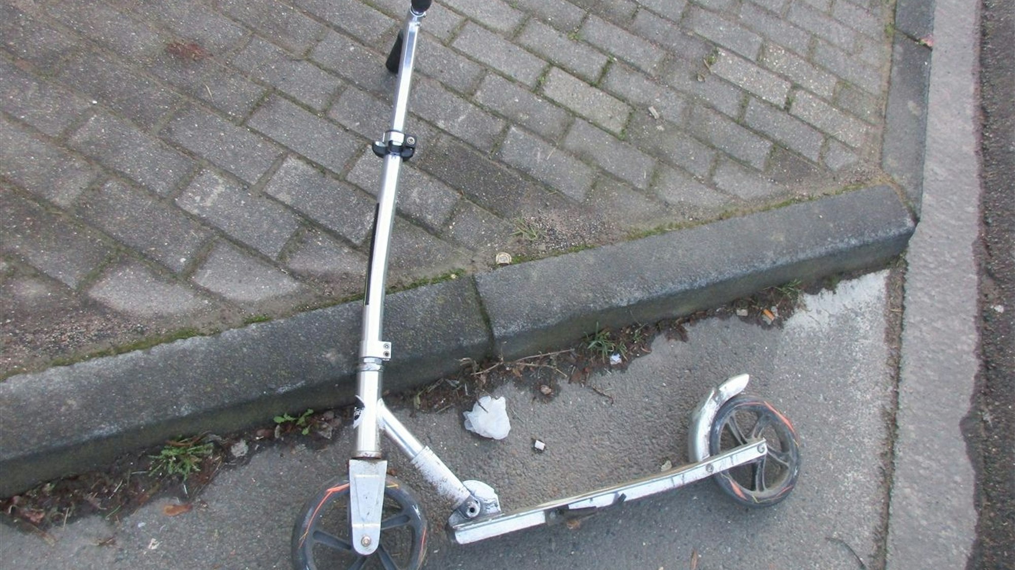 Ein leicht verbogener Roller liegt am Bordstein eines Gehwegs neben einer Straße.