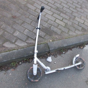 Ein leicht verbogener Roller liegt am Bordstein eines Gehwegs neben einer Straße.