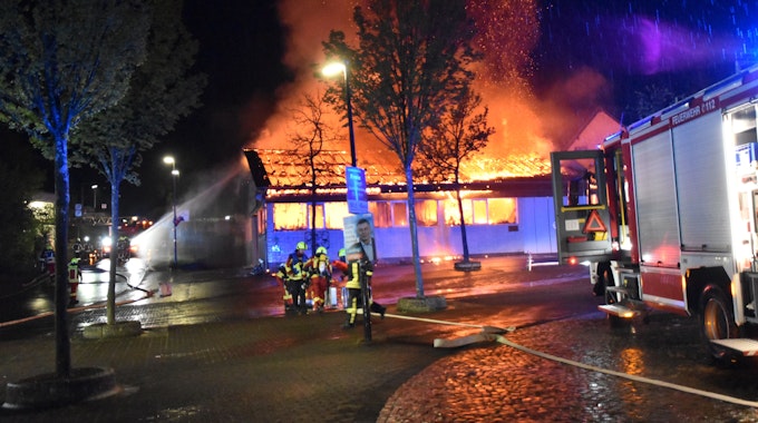 Eine Markthalle steht nachts lichterloh in Flammen. Feuerwehrleute versuchen mit Wasser, die Flammen zu löschen.