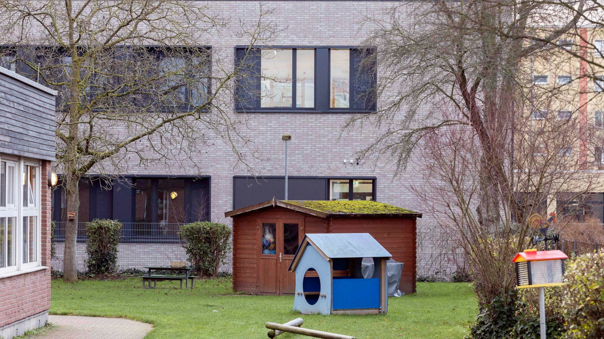 Es ist ein grauer verklinkerter Schulbau zu sehen. Davor ein Rasen mit Spielgeräten und einem blauen Schuppen.