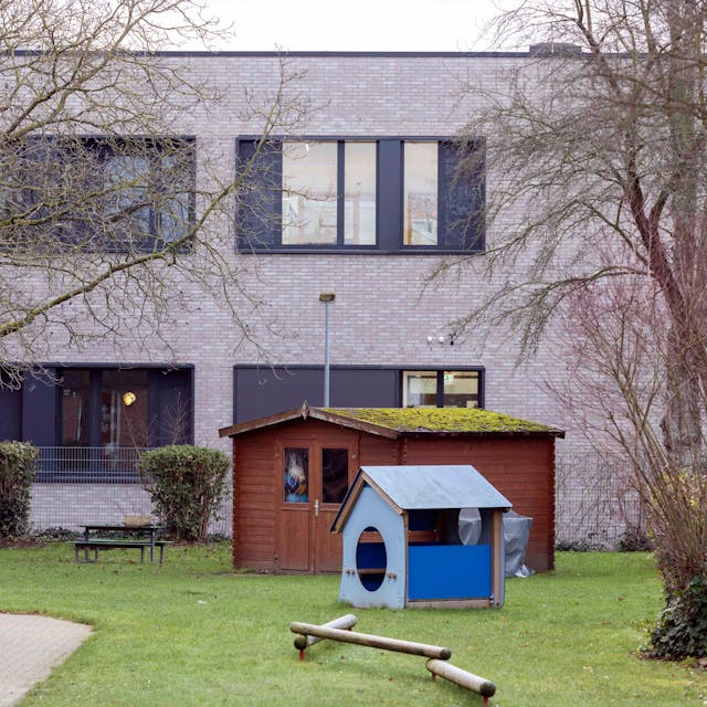 Es ist ein grauer verklinkerter Schulbau zu sehen. Davor ein Rasen mit Spielgeräten und einem blauen Schuppen.