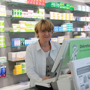 Eine Frau mit blonden Haaren und Brille im weißen Kittel steht in einer Apotheke.