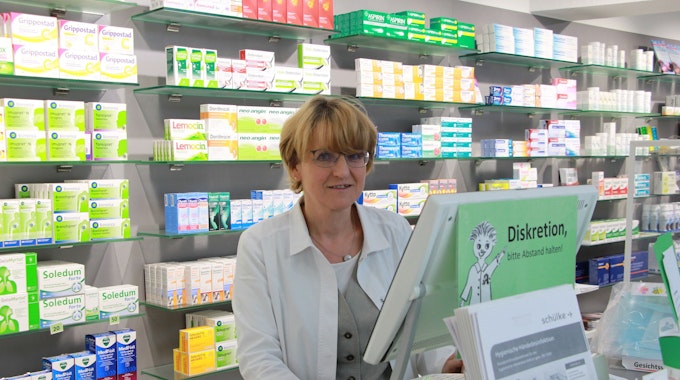Eine Frau mit blonden Haaren und Brille im weißen Kittel steht in einer Apotheke.