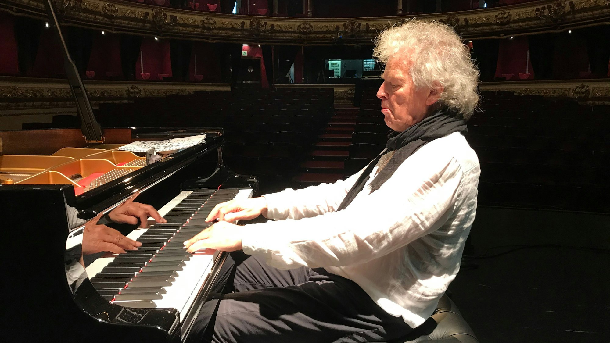 Der Jazz-Musiker Joachim Kühn sitzt in einem leeren Konzertsaal am Klavier. Das Licht ist auf ihn gerichtet und er trägt ein weißes Leinenhemd mit schwarzem Schal.