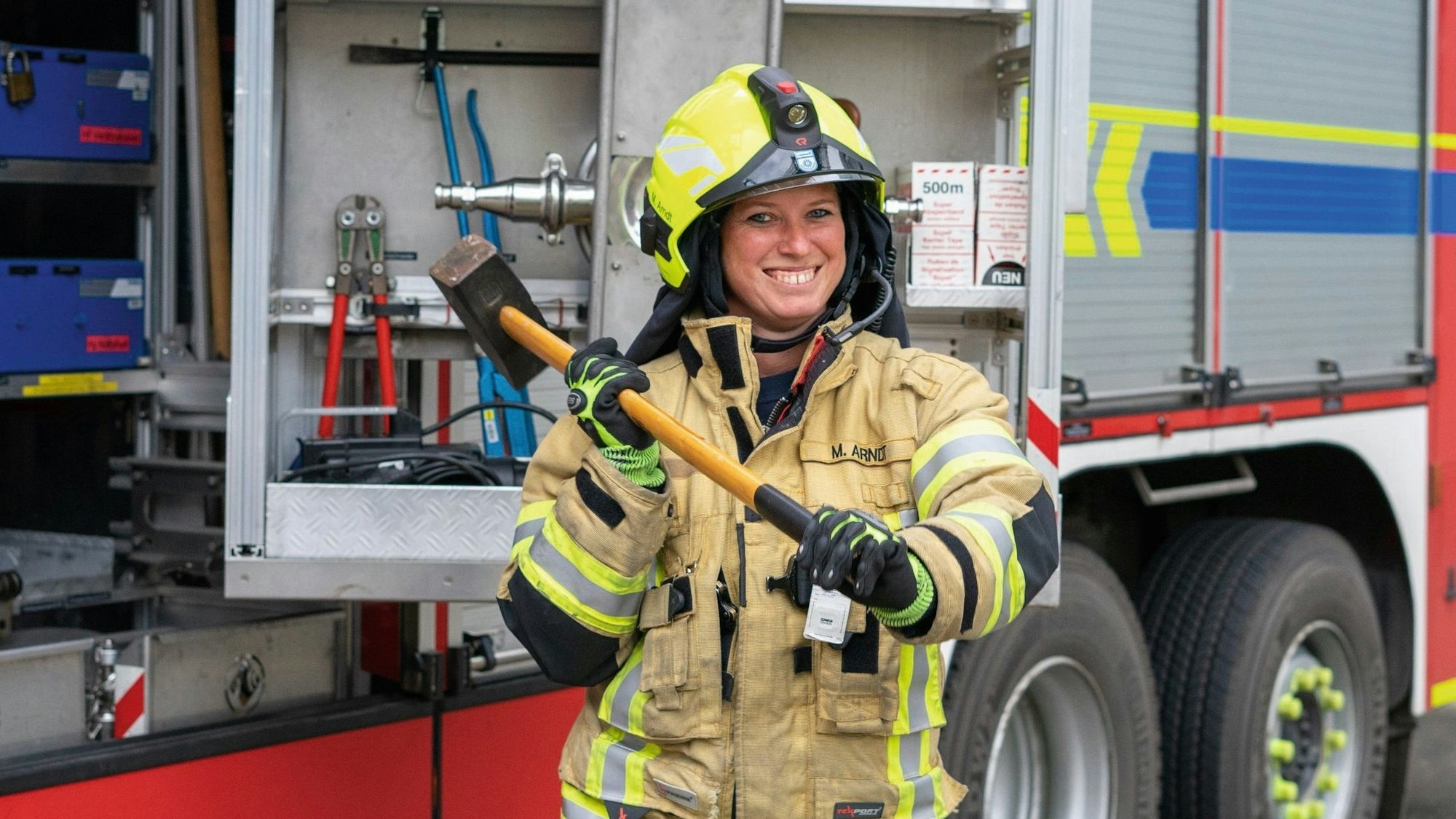 Eine Feuerwehrfrau steht in Einsatzkleidung vor einem Feuerwehrwagen, lächelt in die Kamera und schwingt einen Vorschlaghammer.
