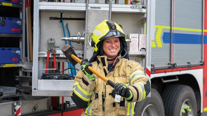 Eine Feuerwehrfrau steht in Einsatzkleidung vor einem Feuerwehrwagen, lächelt in die Kamera und schwingt einen Vorschlaghammer.