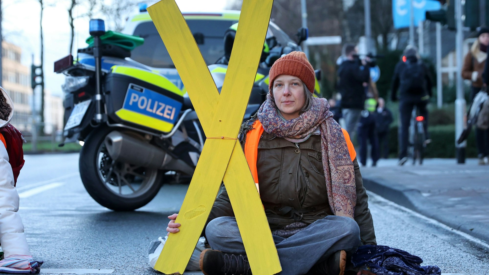 Caroline Schmidt von der Letzten Generation sitzt auf einer Straße im Schneidersitz. Eine Hand ist auf dem Boden angeklebt. In der anderen Hand trägt sie ein gelbes X.