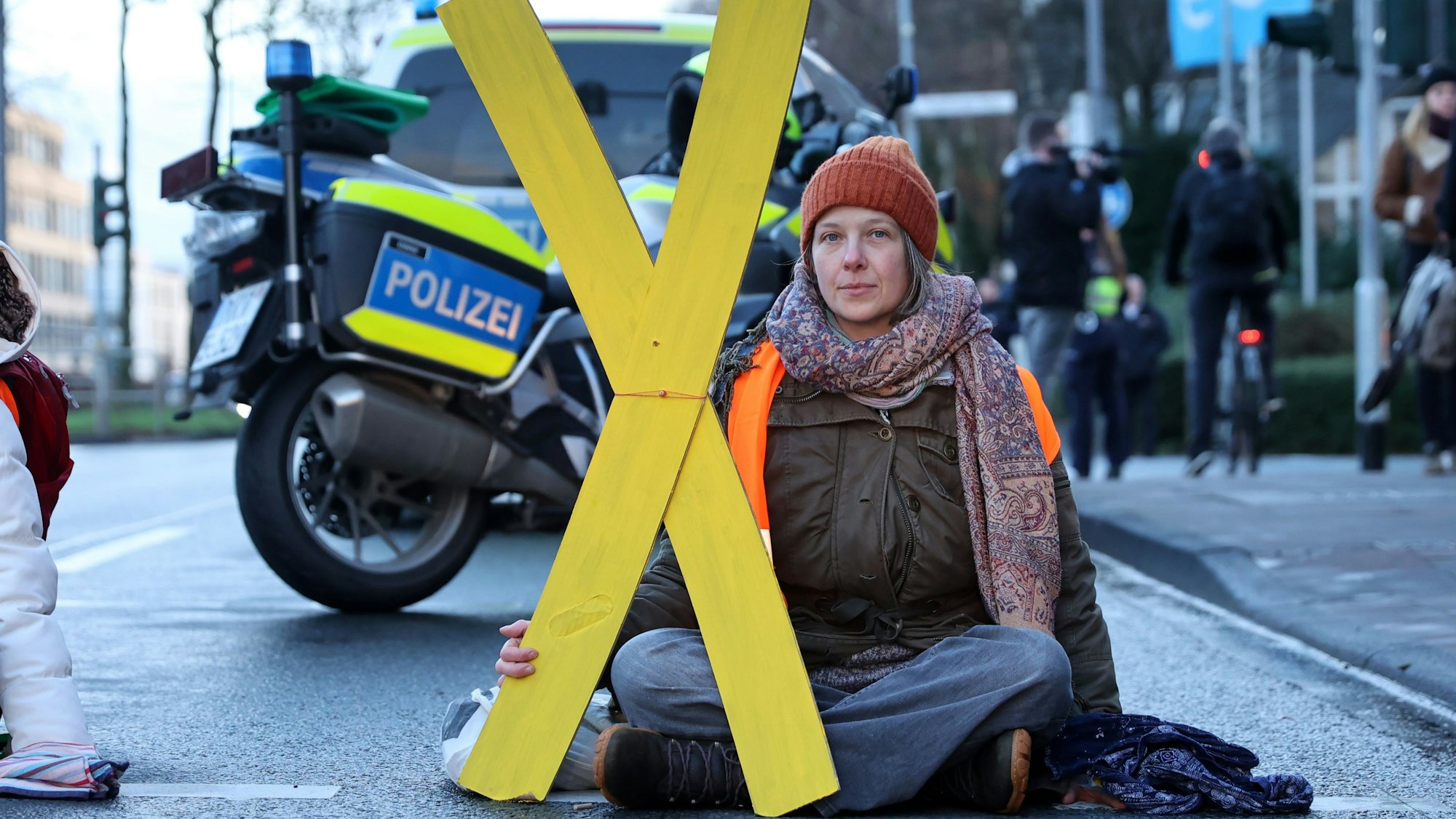 Caroline Schmidt hat ein X aus Holz in der Hand und sitzt auf der Straße, hinter ihr steht ein Motorrad der Polizei.