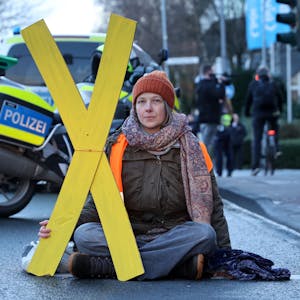 Caroline Schmidt von der Letzten Generation sitzt auf einer Straße im Schneidersitz. Eine Hand ist auf dem Boden angeklebt. In der anderen Hand trägt sie ein gelbes X.&nbsp;